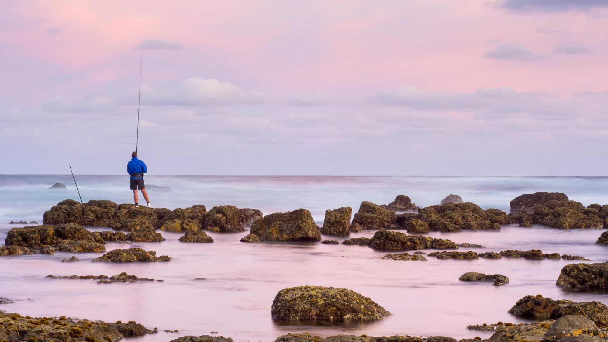 A man in a blue jacket stands on dark, jagged rocks along the shoreline, fishing at sunset as pastel hues of pink and lavender blend across the cloudy sky.
