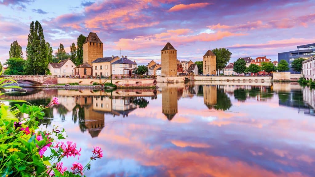 Pastel-colored sunset over Strasbourg, France, with reflections of medieval towers and houses in the calm river, bordered by blooming flowers in the foreground.