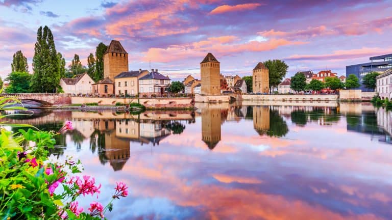 Pastel-colored sunset over Strasbourg, France, with reflections of medieval towers and houses in the calm river, bordered by blooming flowers in the foreground.