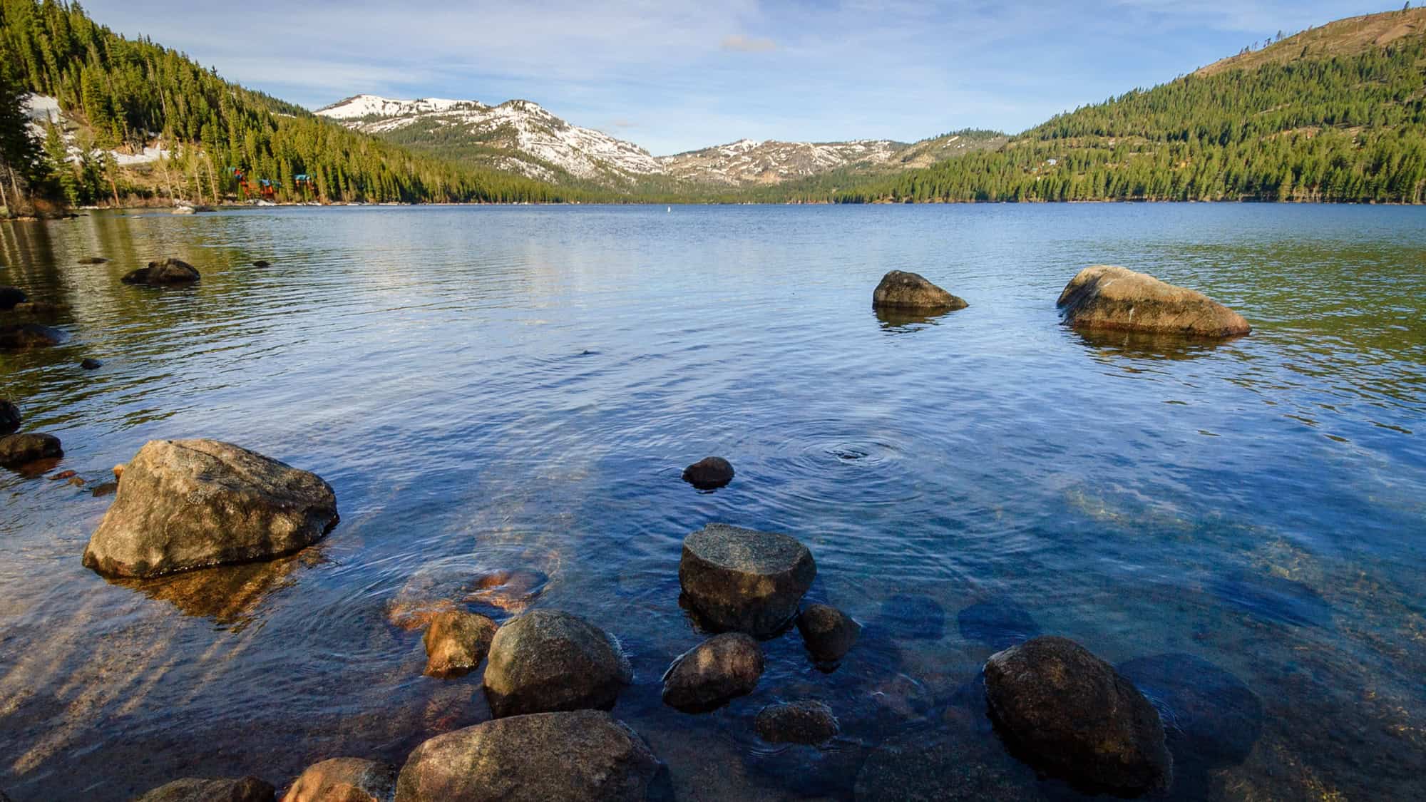 A serene view of Donner Lake with large rocks in the clear foreground water, pine-covered hills on both sides, and snow-capped mountains in the distance under a blue sky.