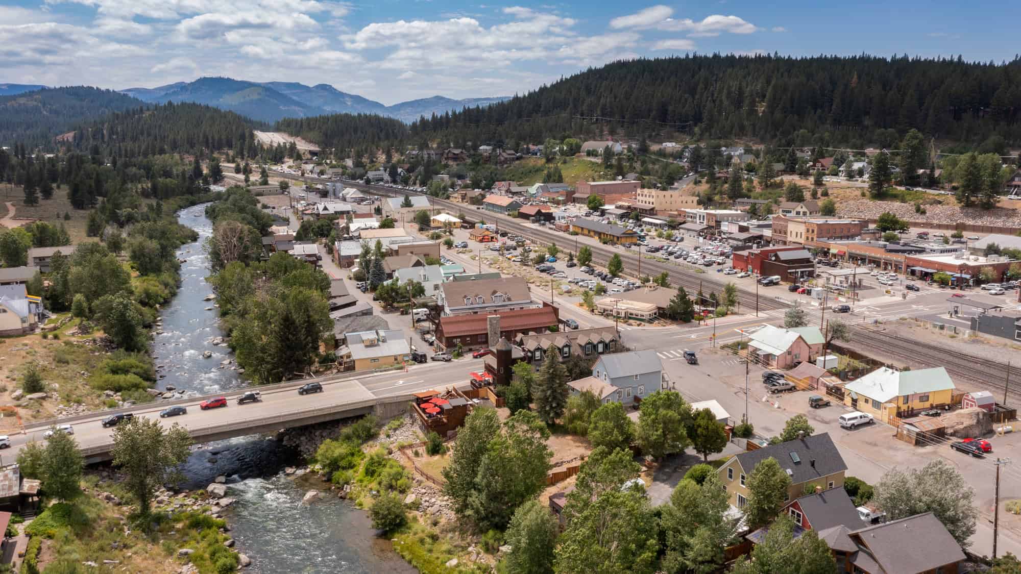 A high-angle view of downtown Truckee, California, showing a river winding through the town, rail lines, clustered buildings, and forested hills beyond.