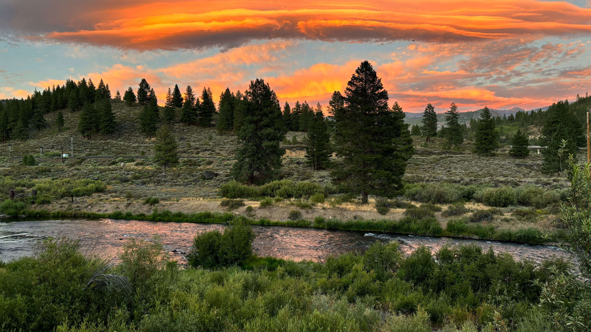 A vibrant sunset over the Truckee River, with fiery orange clouds glowing above a pine-dotted landscape and brushy riverbanks reflecting the sky’s colors.