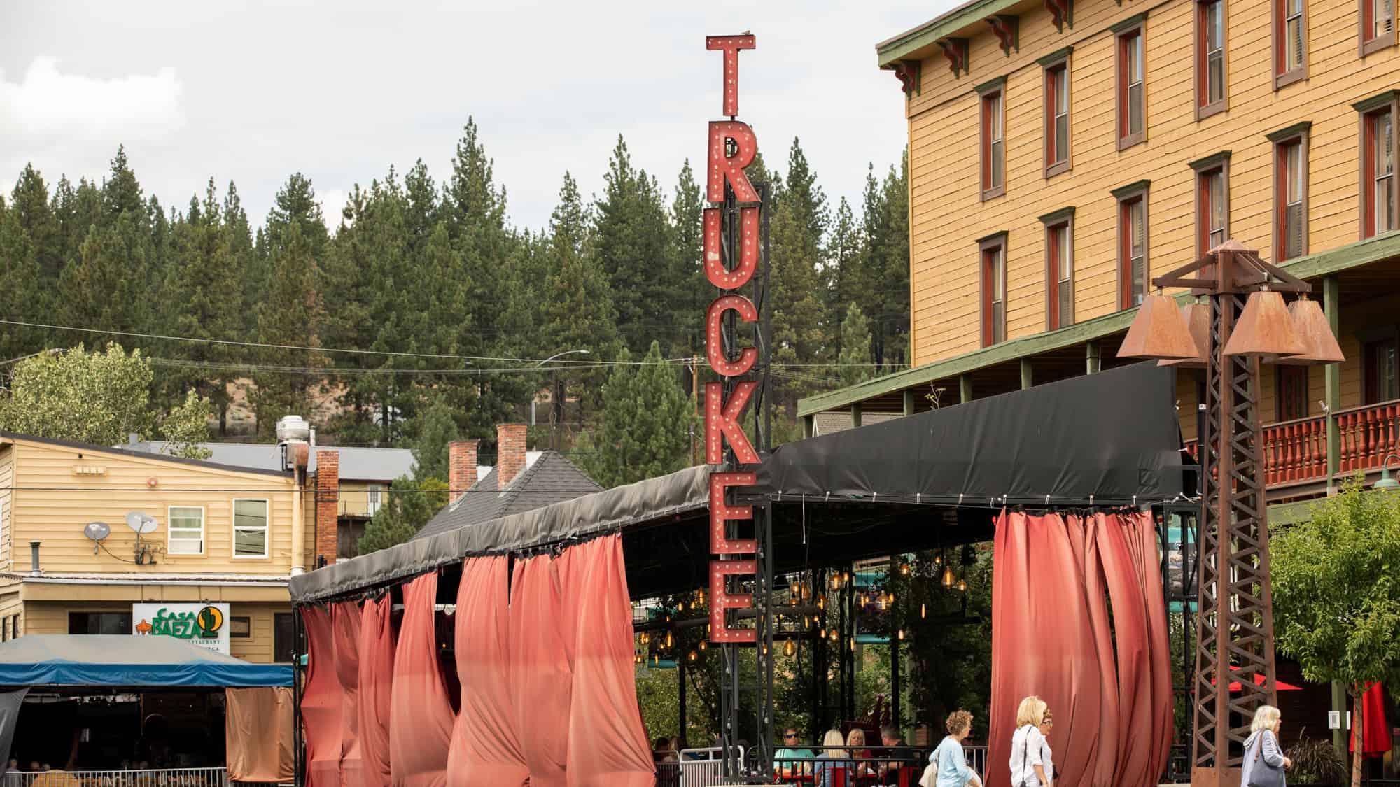 Street-level image of downtown Truckee with a large vertical “TRUCKEE” sign, string-lit outdoor dining space draped in red curtains, and pine trees in the background.