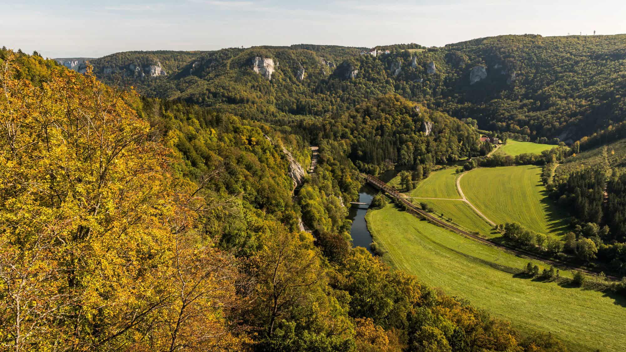 A lush, tree-covered valley with a winding river and trails, surrounded by forested cliffs and dotted with farm buildings and a small bridge, viewed from a high vantage point in golden autumn light.