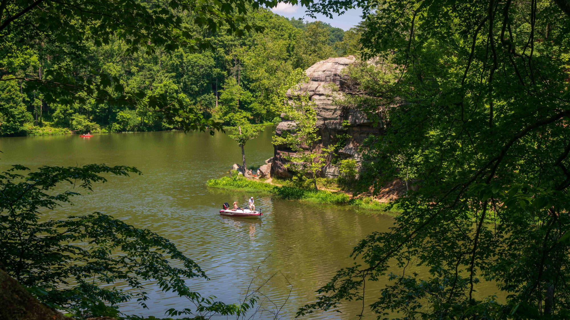 A peaceful lake surrounded by dense, green forest is framed by overhanging branches, with a few people in a small boat enjoying a calm afternoon on the water.