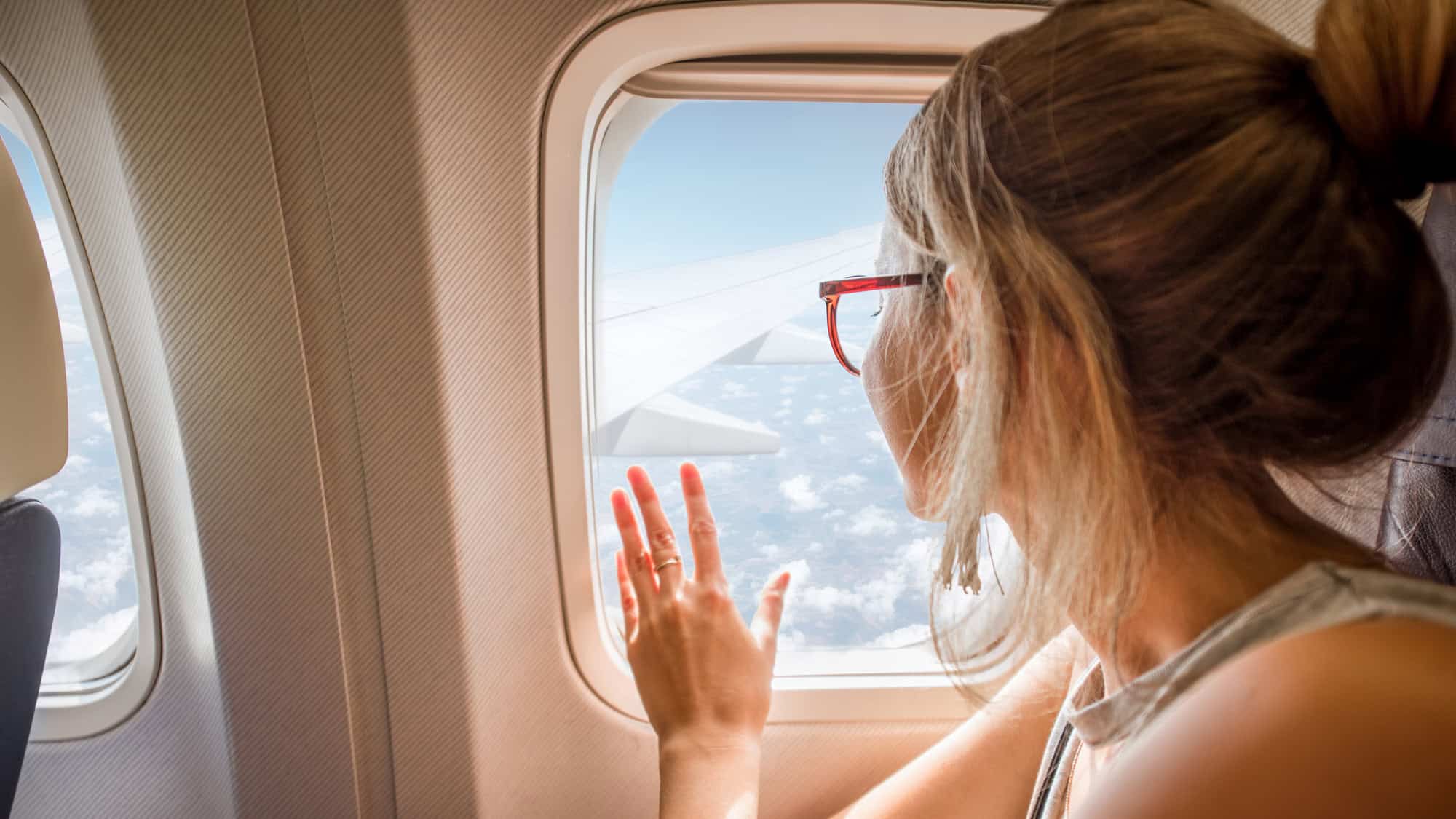 A woman with a messy bun and red glasses gazes out an airplane window, hand gently pressed against the glass as she admires the sky and clouds outside.