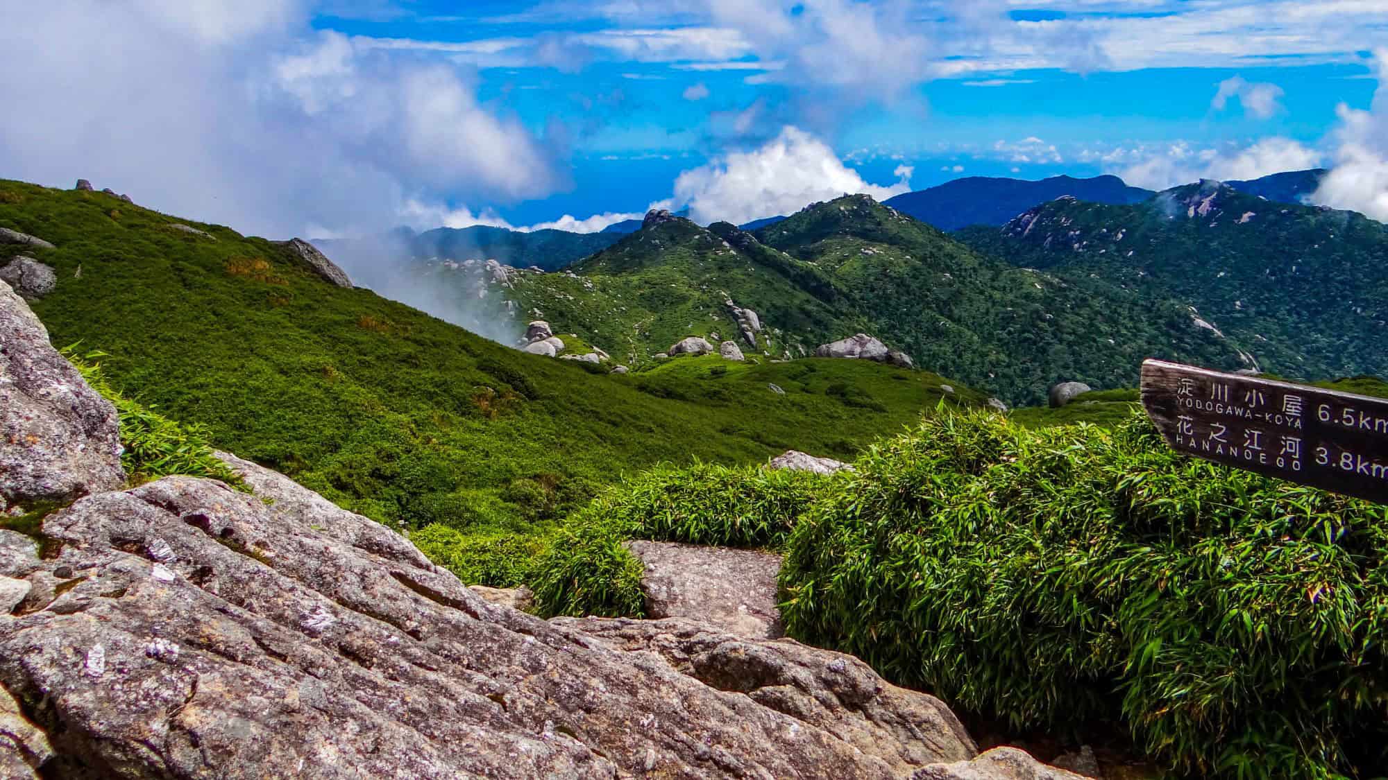 A rocky mountain trail winds through the lush, green hills of Yakushima Island in Japan, with a wooden sign in Japanese and English marking trail distances.