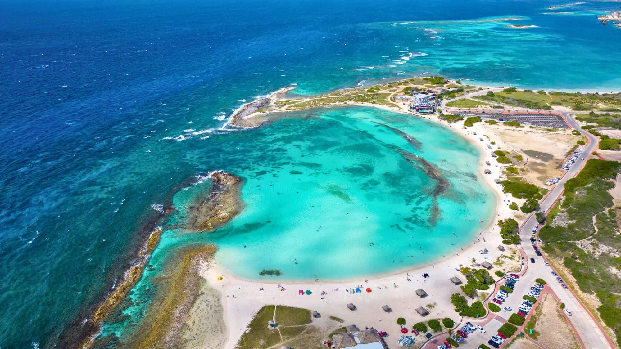 Aerial view of a crescent-shaped beach with bright turquoise waters. The beach is lively with people, umbrellas, and small huts. A parking lot and facilities are visible, indicating a well-developed tourist spot.