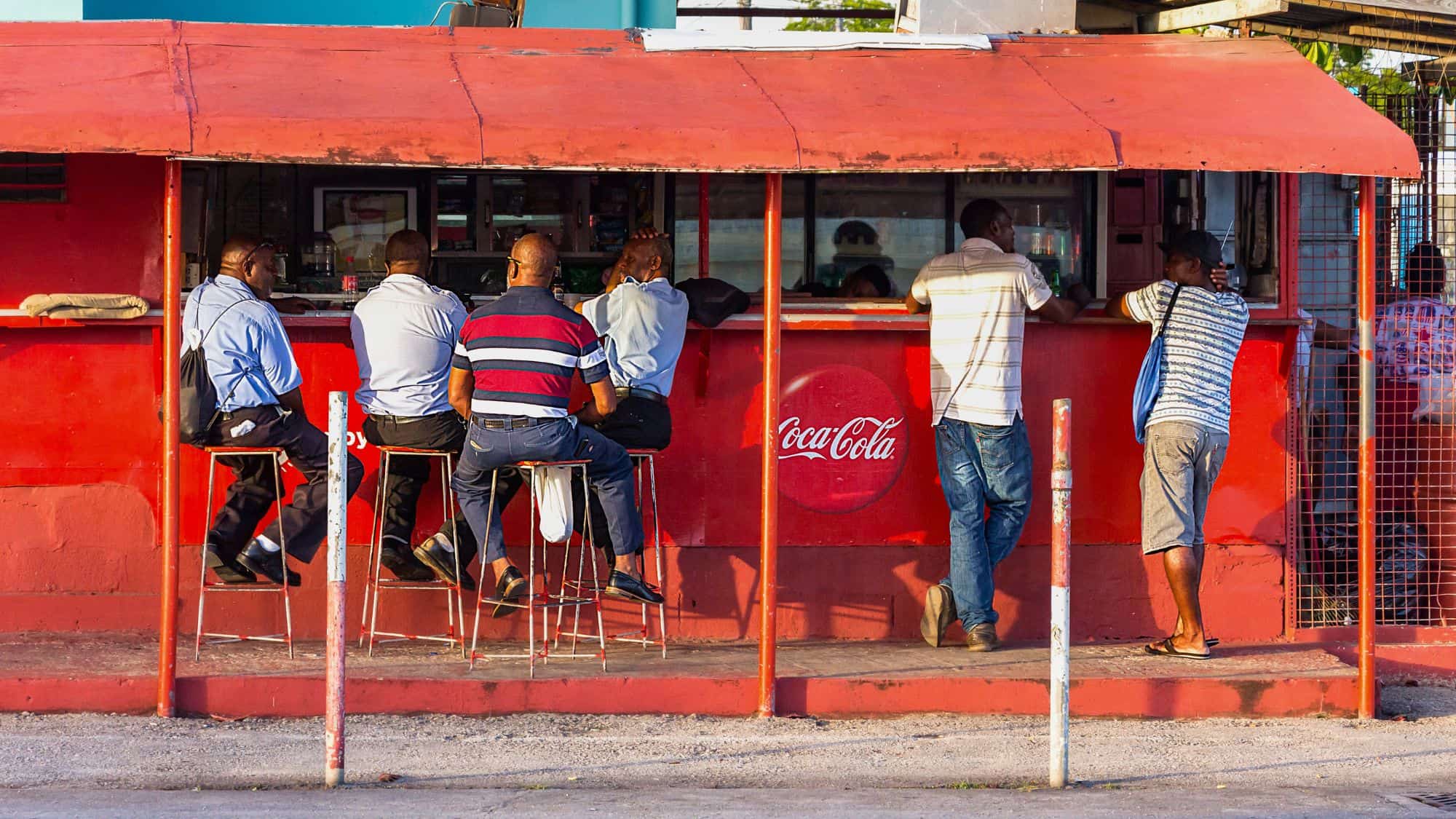 Men relax at a simple red street bar adorned with Coca-Cola branding, sharing conversation and drinks in the late afternoon light.