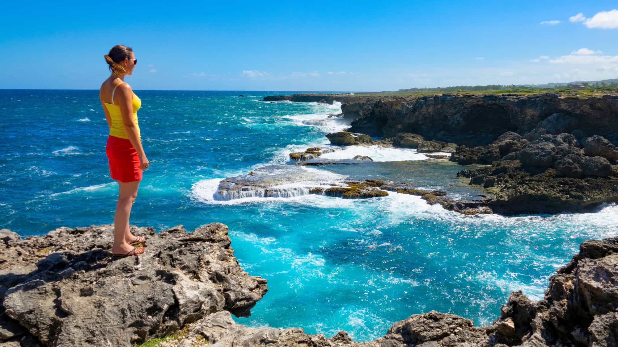 A woman in a yellow top and red shorts stands on a cliffside overlooking the vibrant turquoise surf crashing against the rugged northeastern coast of Barbados.