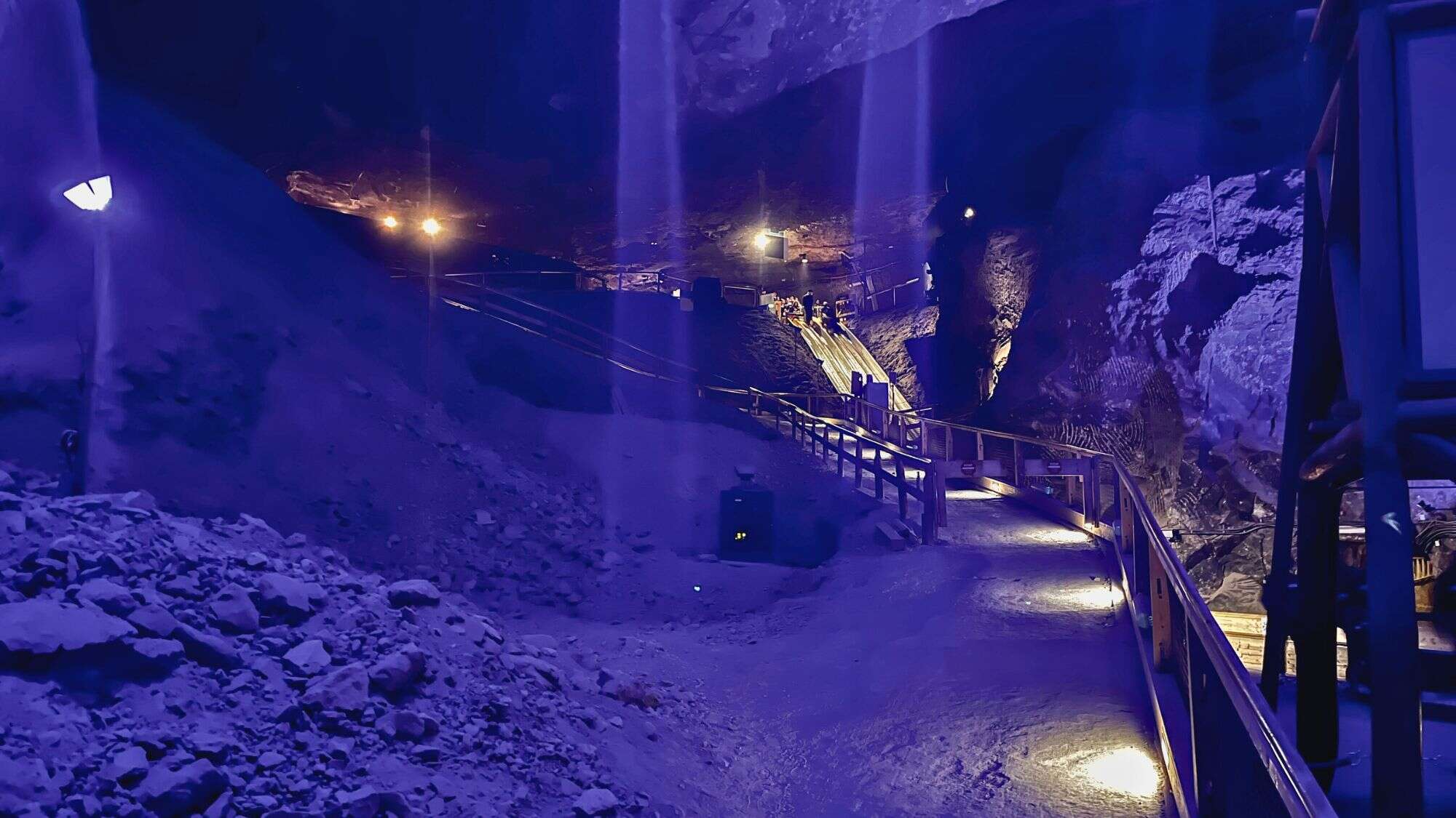 An underground salt cavern is bathed in dramatic blue lighting, highlighting a sloped path with handrails and embedded floor lights that lead toward a distant illuminated slide structure.
