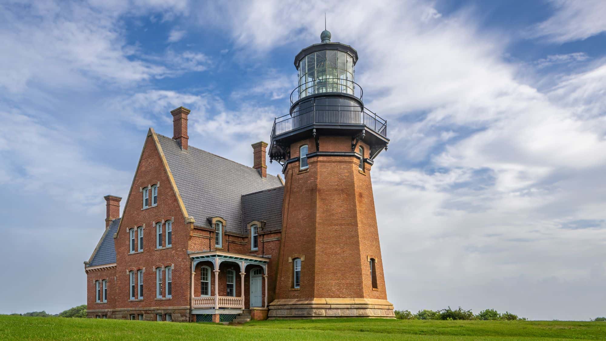 A stately brick lighthouse with Victorian architecture and a black lantern tower stands on a grassy bluff under a partly cloudy blue sky.