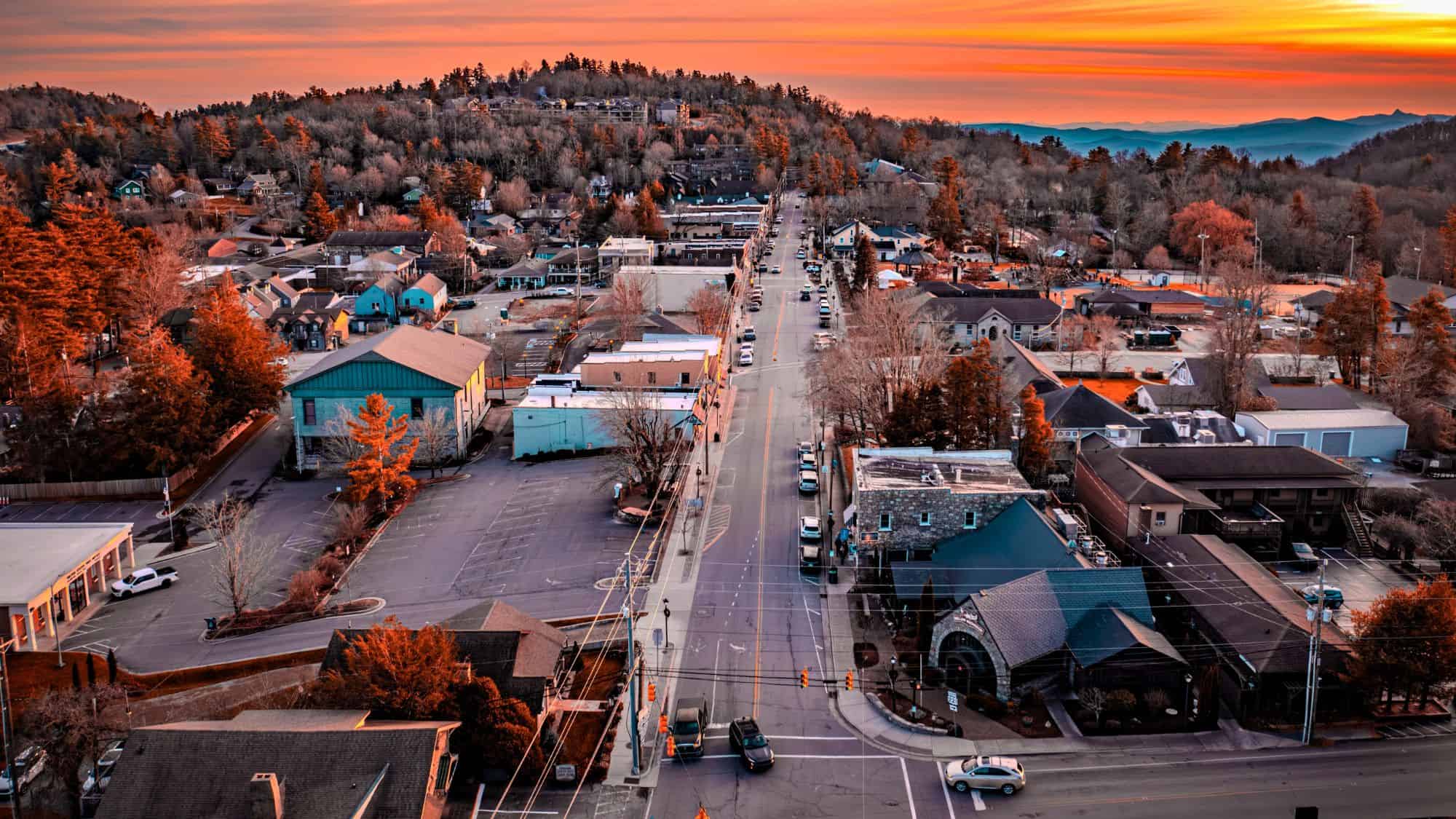 A vibrant sunset bathes a quaint mountain town in warm light, highlighting rooftops, tree-covered hills, and a straight main road lined with cars and shops.