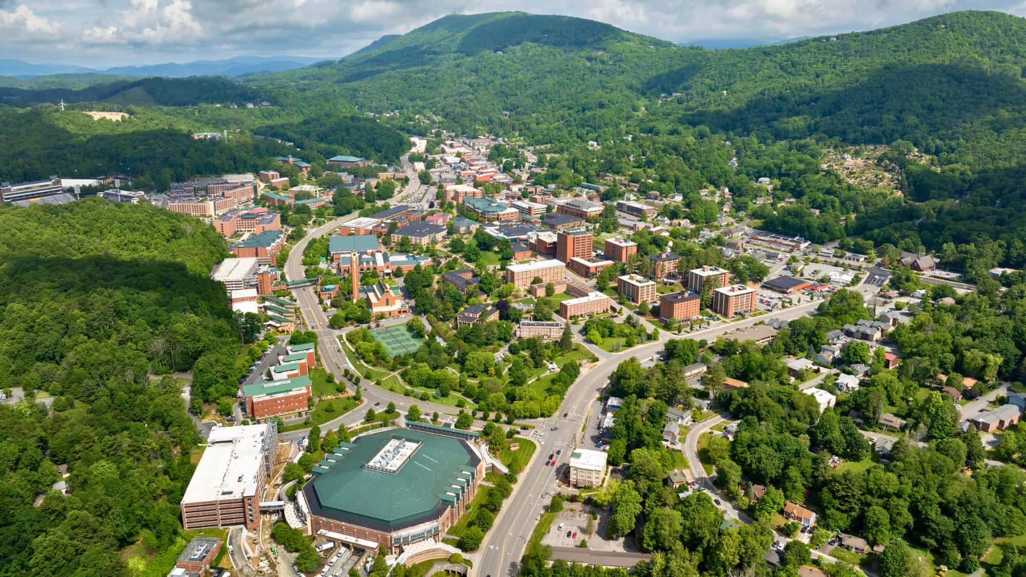 An aerial view of a small, vibrant mountain town surrounded by rolling green hills, with a mix of red-brick buildings, residential homes, and winding roads nestled in dense forest.