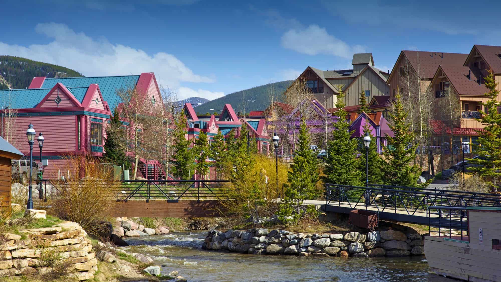 A small alpine town features brightly painted buildings in pink, purple, and red tones nestled among evergreens and crossed by a creek, with forested hills rising behind. Ask ChatGPT