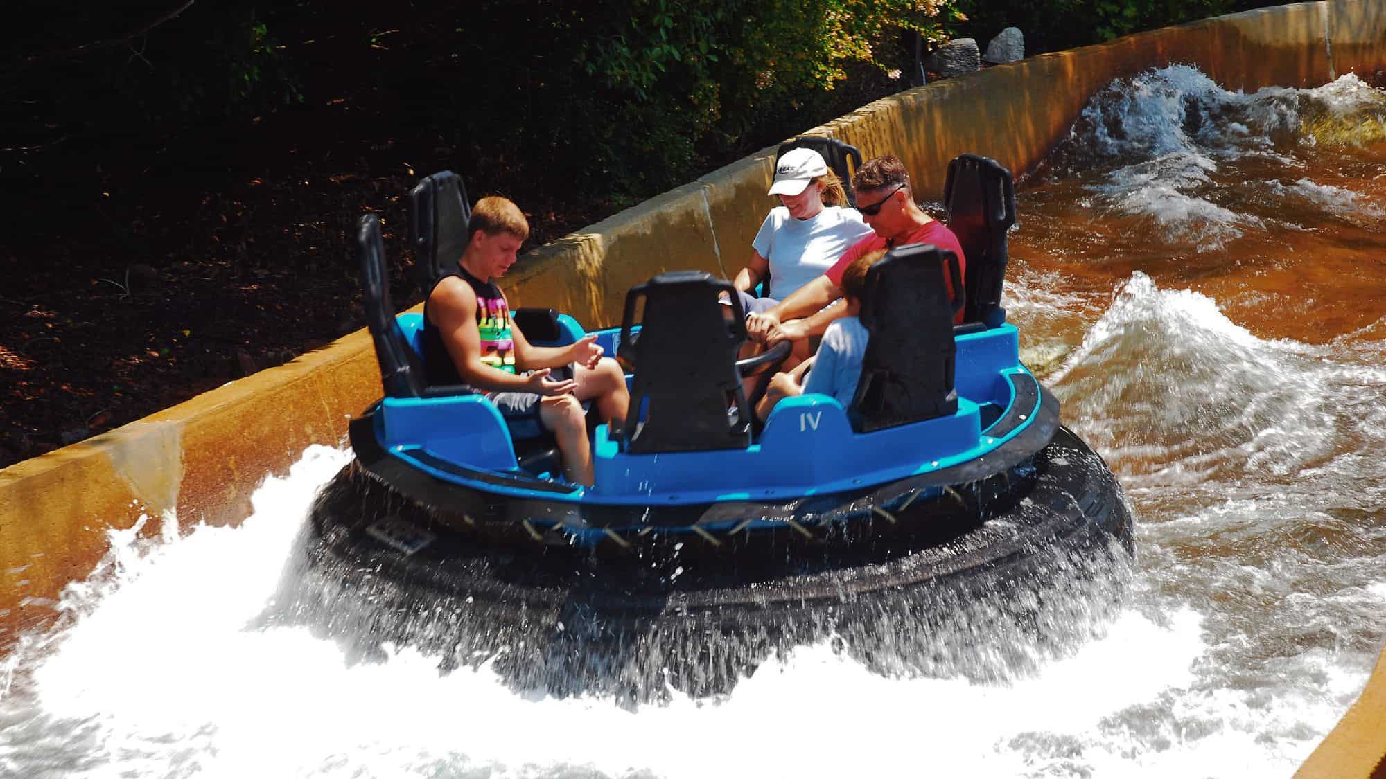 A group of people ride a circular blue raft through fast-moving water on a whitewater rapids attraction, with water splashing up around them.