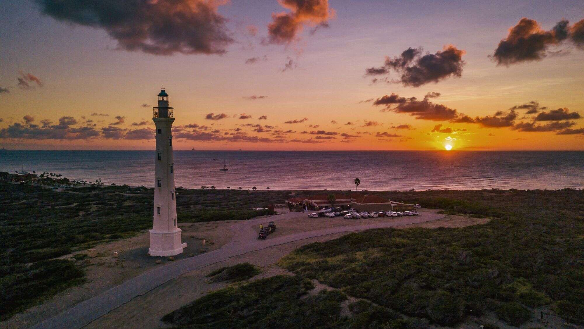 A serene coastal scene featuring a tall white lighthouse against a vibrant sunset sky. The ocean is calm, with small boats in the distance, and the foreground includes sandy paths and low vegetation.
