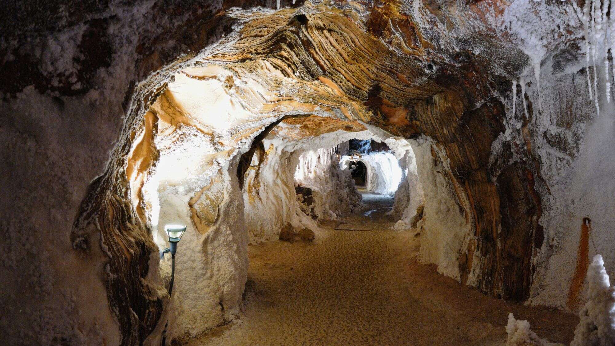 A winding underground tunnel lined with thick salt deposits and layered rock formations, softly illuminated by ground lighting.
