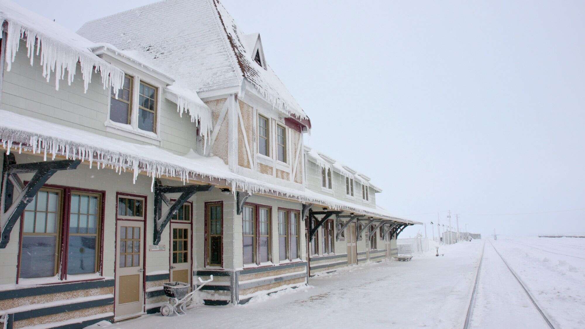 A snow-covered train station with icicles hanging from the roof sits beside empty tracks, surrounded by a stark, wintry white landscape.
