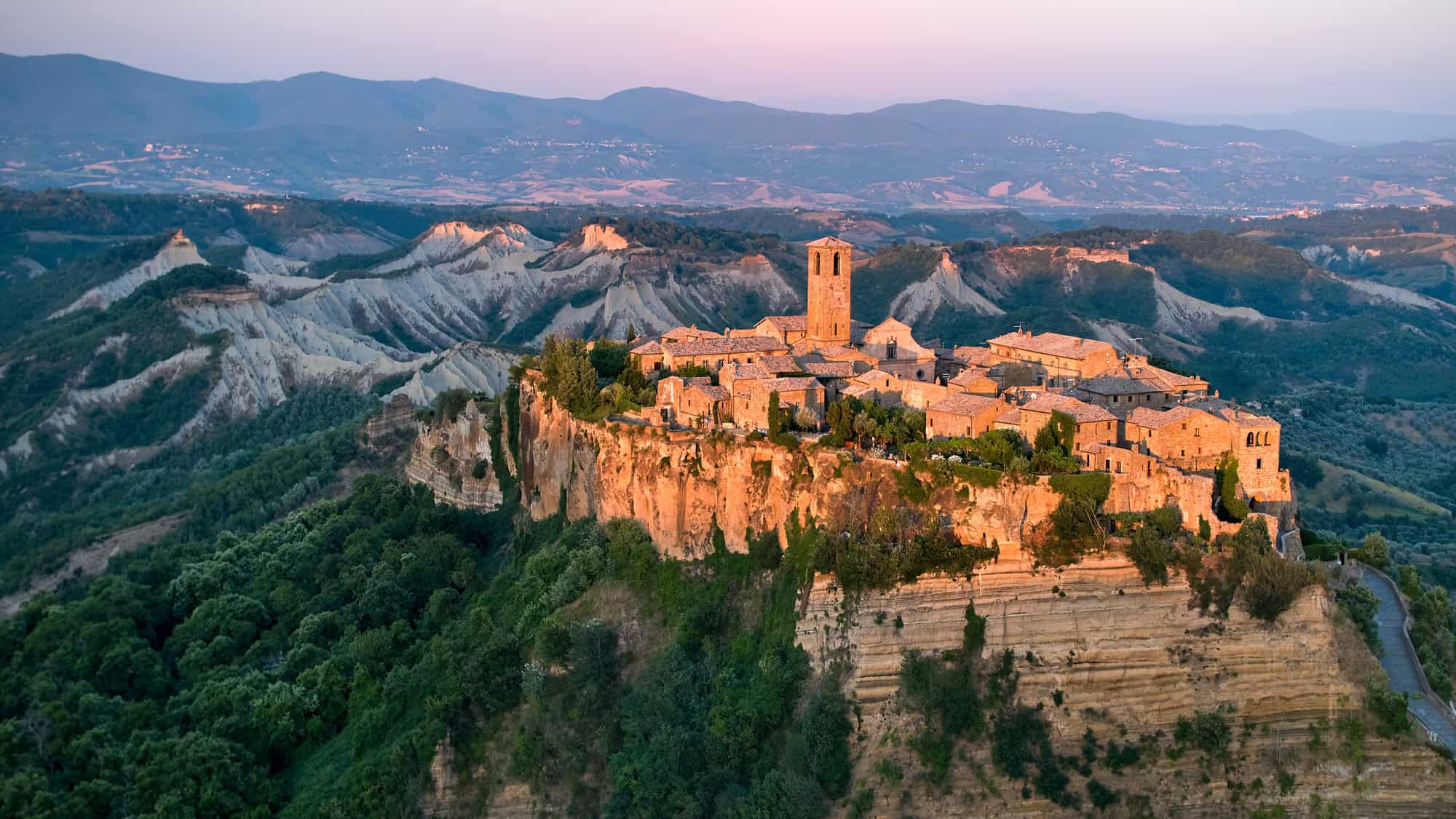 The medieval town of Civita di Bagnoregio in Italy is dramatically perched atop a cliff, glowing in the golden hour light and surrounded by rugged hills and deep green valleys.
