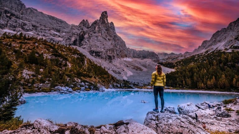 A hiker in a yellow jacket stands on a rock by a turquoise lake, watching the dramatic sunset illuminate jagged mountains in shades of pink and orange.