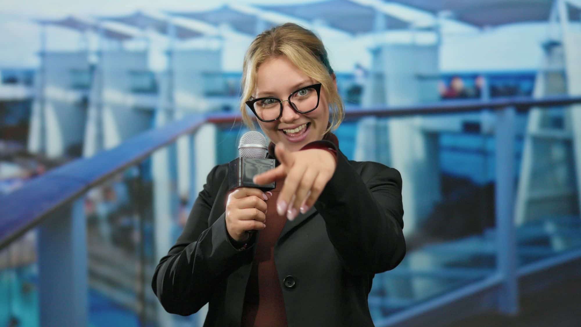 A smiling woman with glasses and a microphone points at the camera, likely an energetic cruise ship host or entertainer engaging guests with a fun announcement or show.