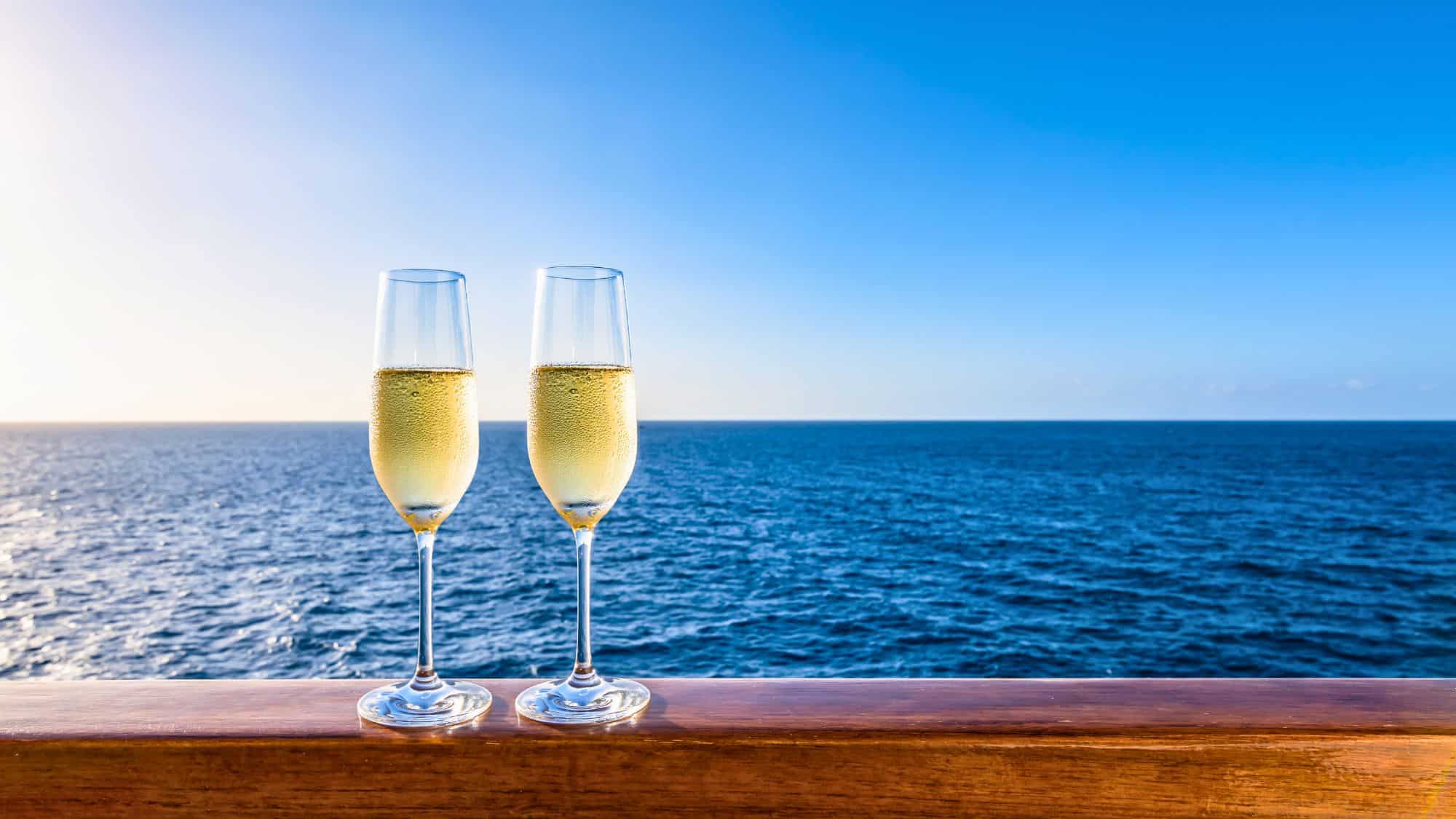 Two flutes of champagne rest on a cruise ship’s railing with the open ocean and a clear blue sky stretching into the distance.