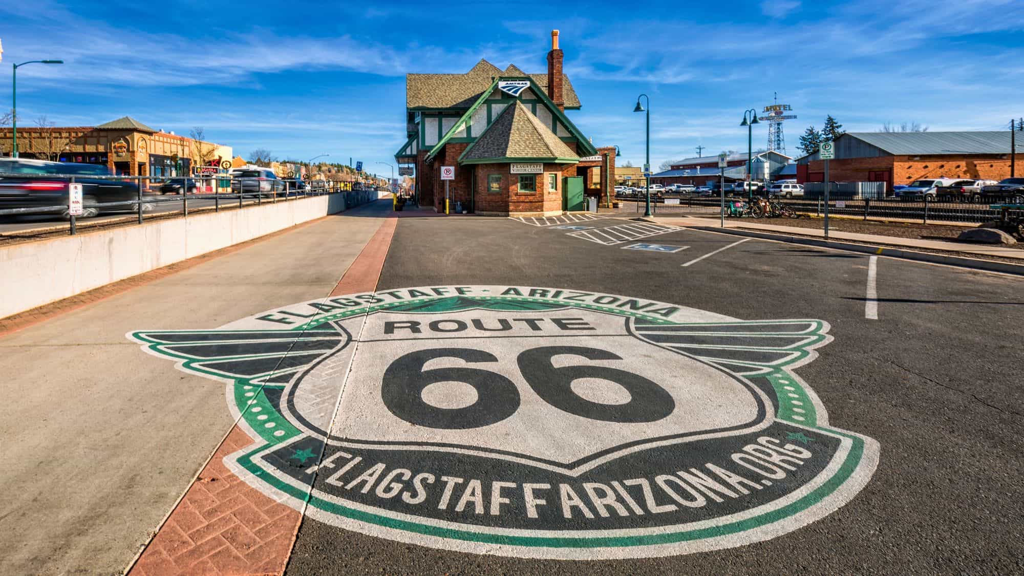 A wide-angle street view in Flagstaff, Arizona features a bold Route 66 emblem painted on the road in front of a historic-looking Amtrak station.