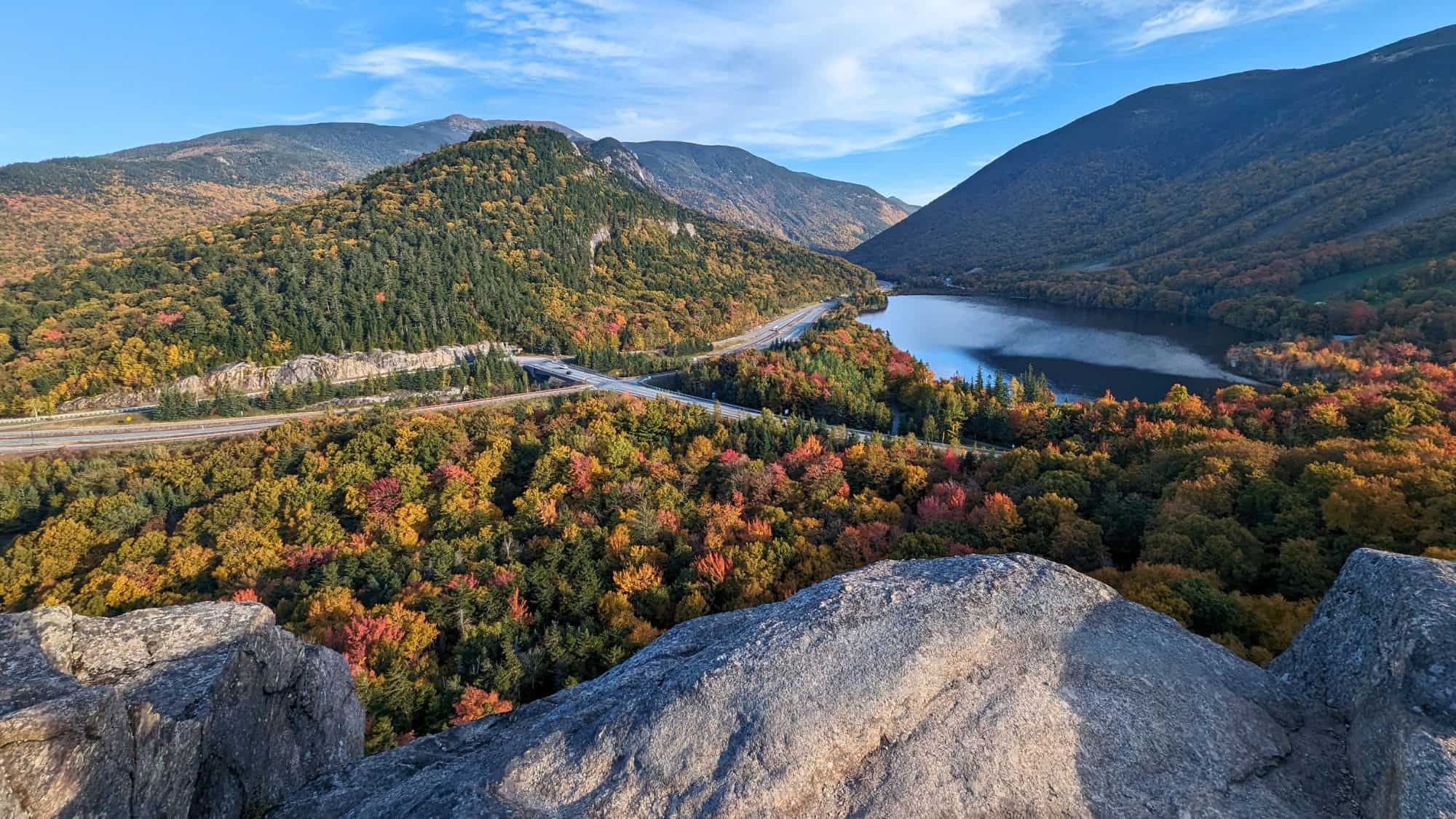 Overlooking a curving highway and reflective lake, this scene captures New Hampshire’s Franconia Notch framed by rocky ledges and vibrant fall foliage.