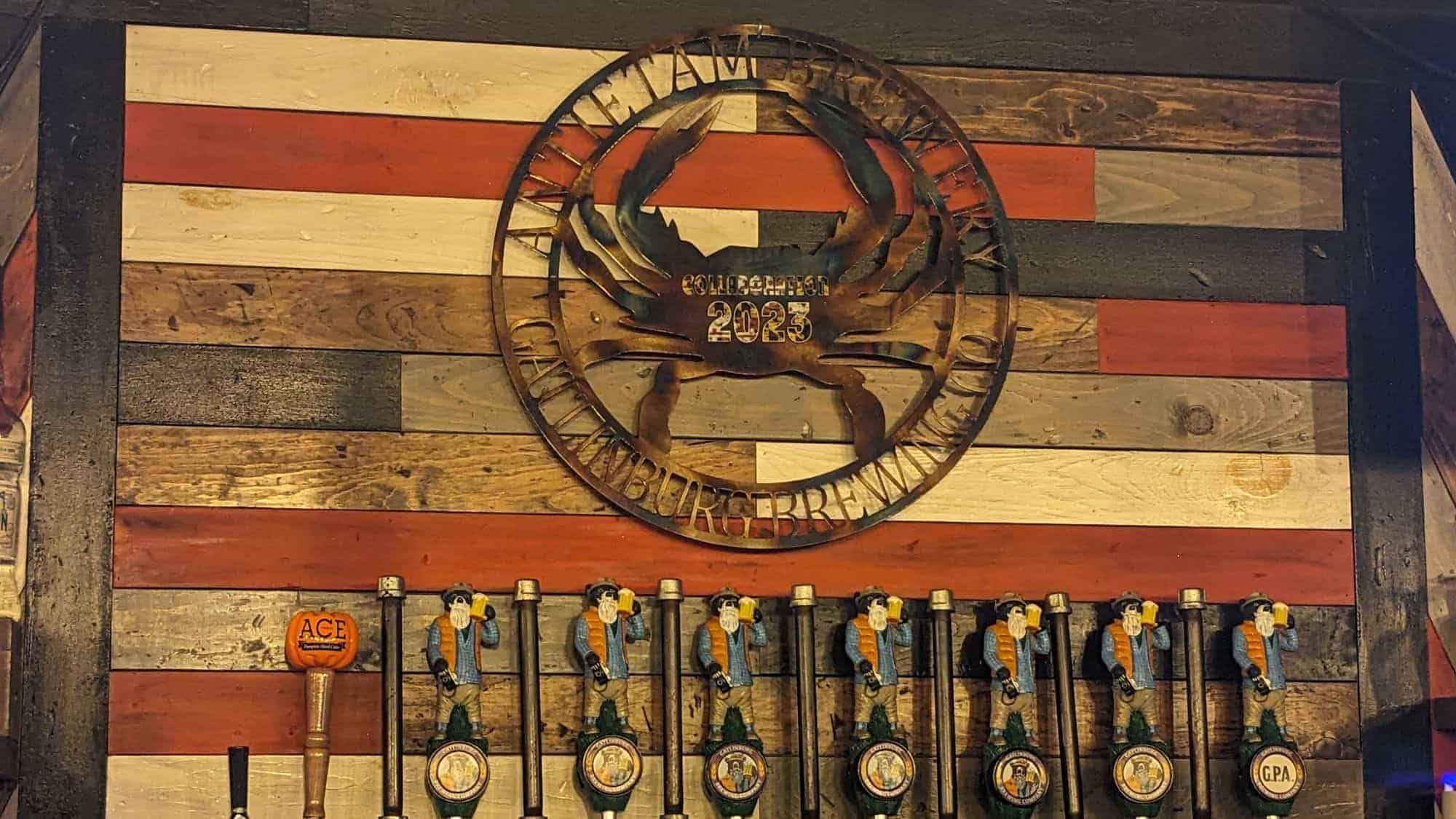 A rustic tap wall with a wooden plank backdrop painted in red, white, and natural wood tones. Above the taps, a circular metal sign reads "Antietam Brewery Collection 2023" with a crab design. The tap handles are shaped like bearded men holding beer mugs, and one tap has an "ACE" pumpkin cider handle.