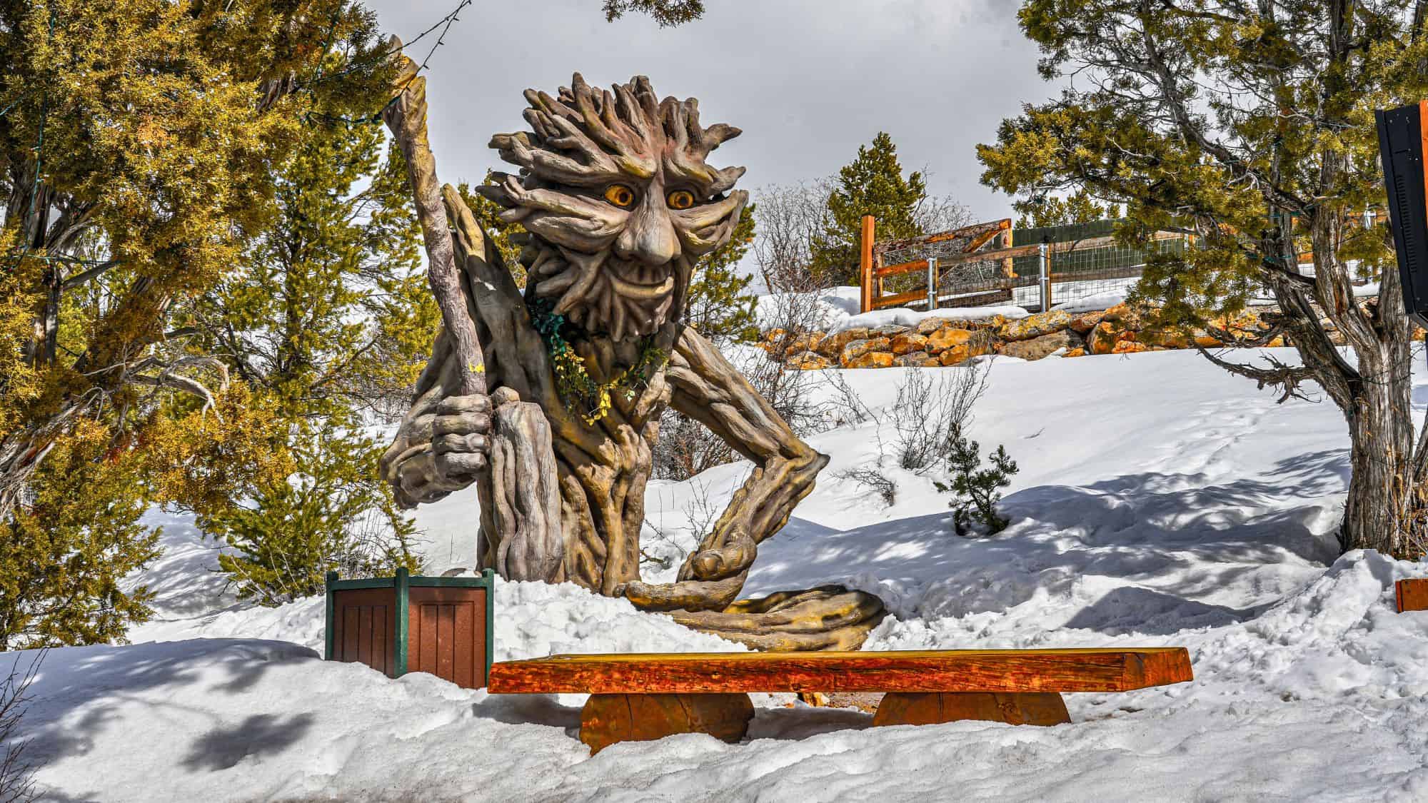 A whimsical wooden tree troll sculpture with yellow eyes and a gnarled staff sits surrounded by snow and evergreen trees.