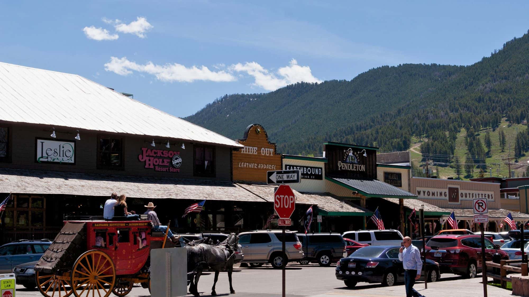 Shops with rustic facades and a horse-drawn stagecoach line a busy street in Jackson Hole, Wyoming, with green hills and ski runs rising behind.