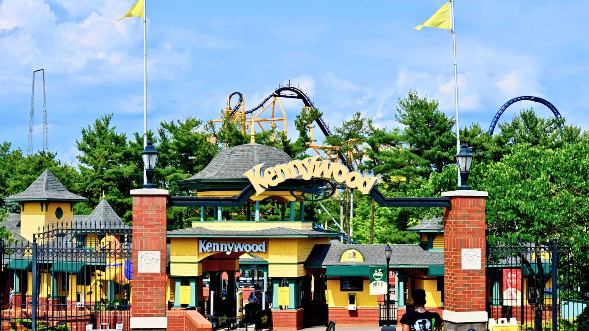 The entrance to Kennywood amusement park features a yellow and green gate with roller coasters rising in the background and flags flying overhead.