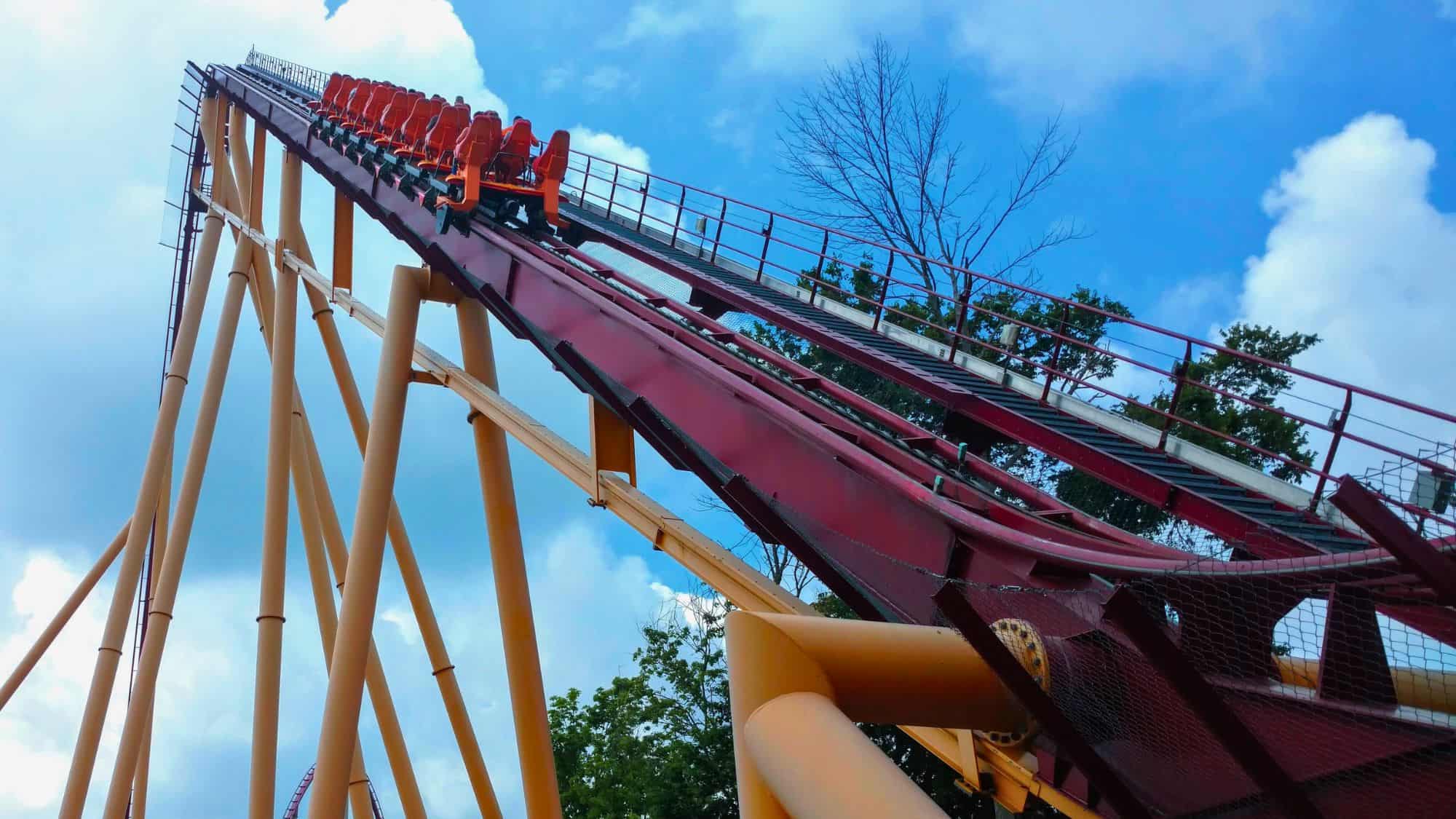 A steep red and orange roller coaster track ascends dramatically into the sky, with riders near the top of the climb.