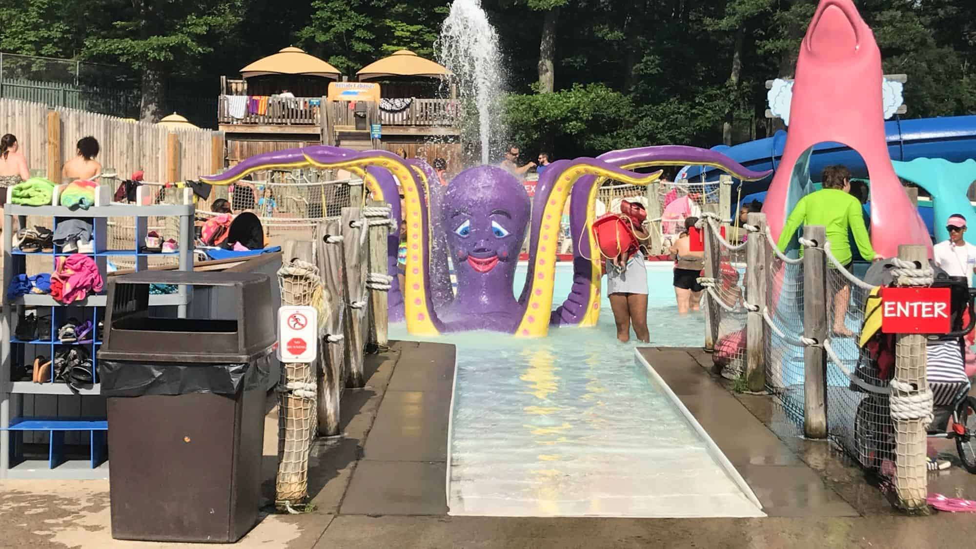 A cheerful purple octopus water feature with yellow tentacles sprays water as kids and lifeguards play around a shallow splash area.