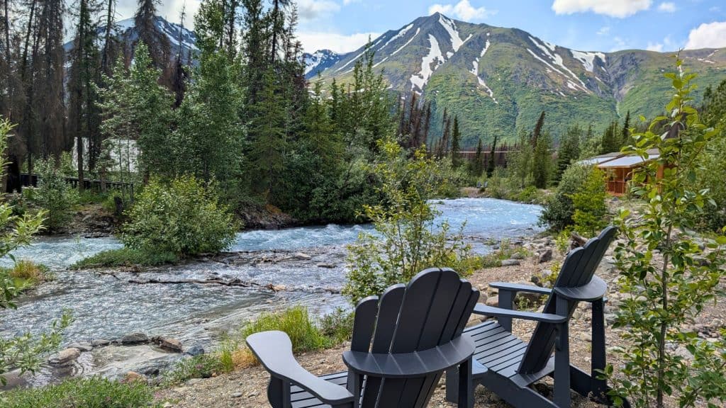 Two black Adirondack chairs sit beside a fast-moving, crystal-clear river surrounded by lush trees and backed by snow-capped mountains under a partly cloudy sky.