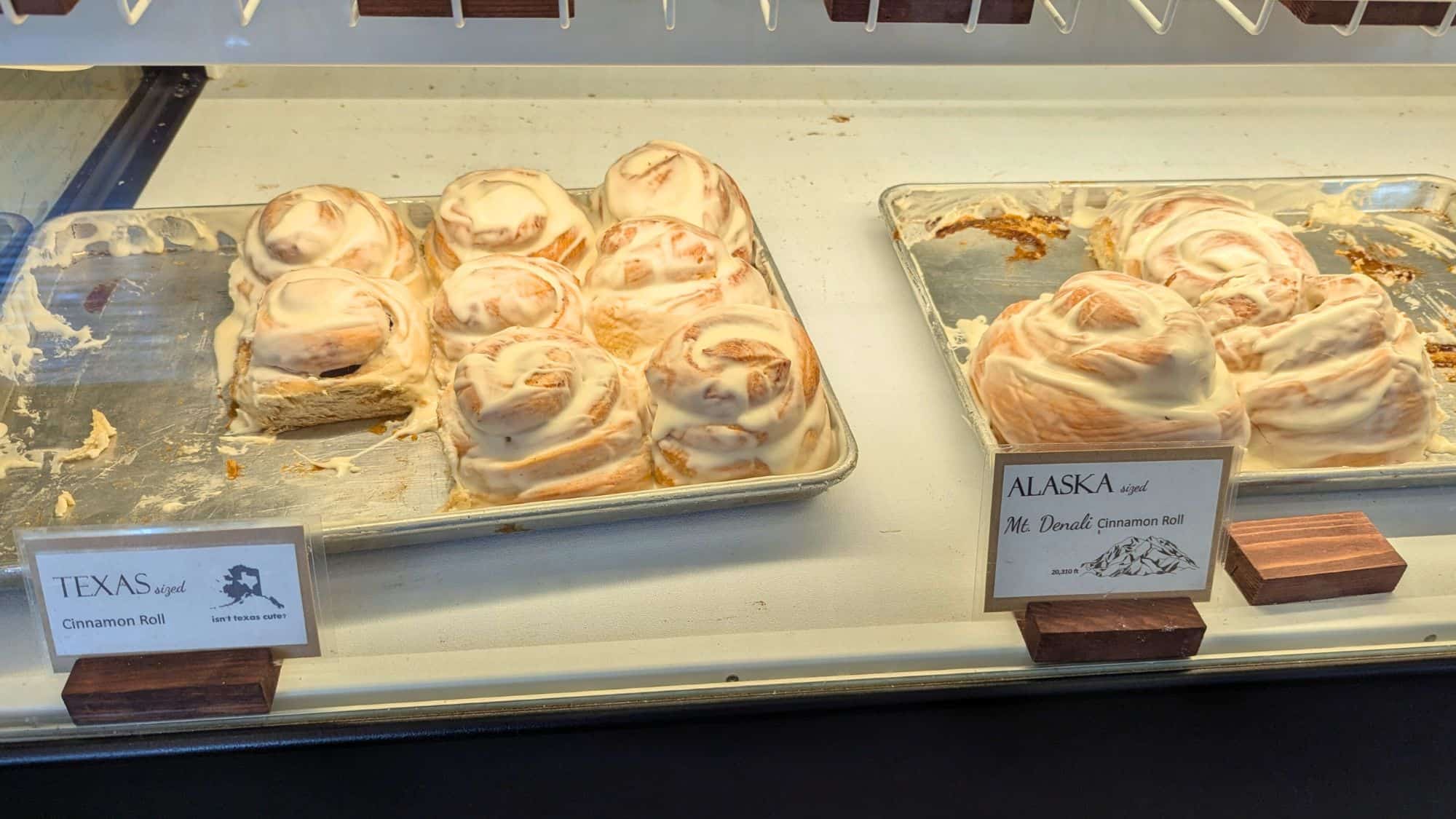 A bakery display features two trays of heavily frosted cinnamon rolls labeled “Texas sized” and “Alaska sized Mt. Denali Cinnamon Roll,” playfully comparing portion sizes.