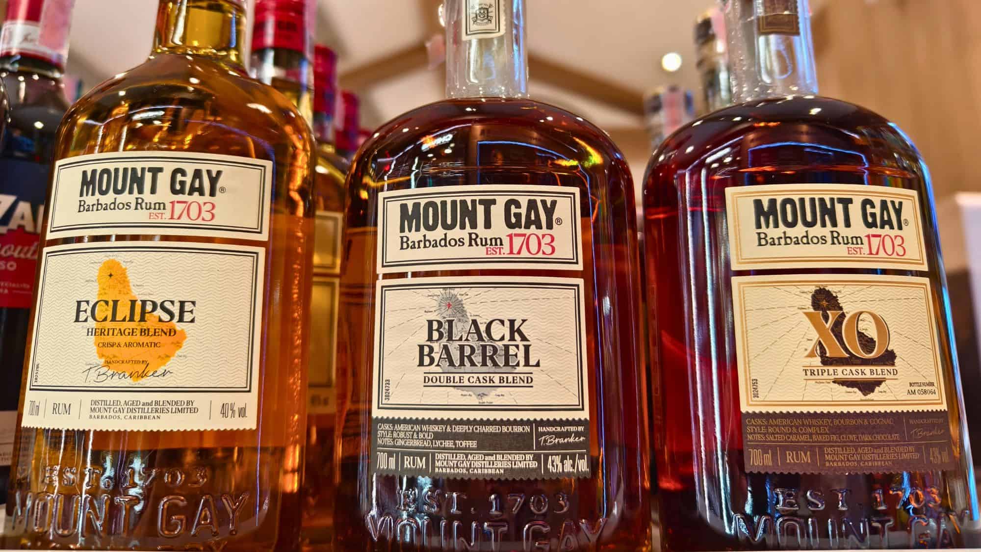 Close-up shot of three bottles of Mount Gay Barbados Rum on a shelf, showcasing the Eclipse Heritage Blend, Black Barrel Double Cask Blend, and XO Triple Cask Blend with detailed labels emphasizing the brand’s Caribbean origin and 1703 establishment.