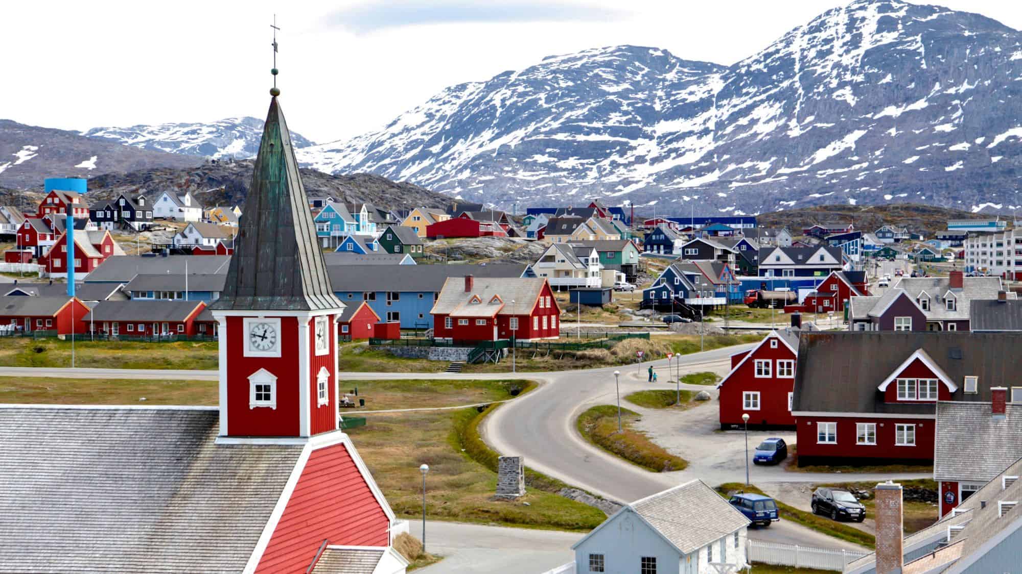A cluster of colorful homes with steep rooftops spreads across the rocky hills of Nuuk, Greenland, with snow-dusted mountains rising behind the town’s red church steeple.
