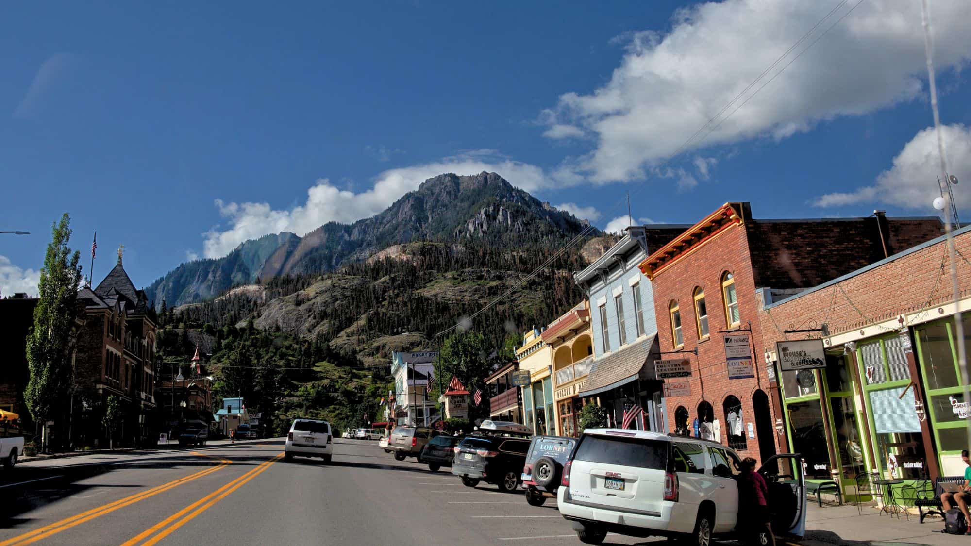 Victorian buildings and modern cars line a charming main street in Ouray, Colorado, with steep forested mountains towering dramatically above the town.