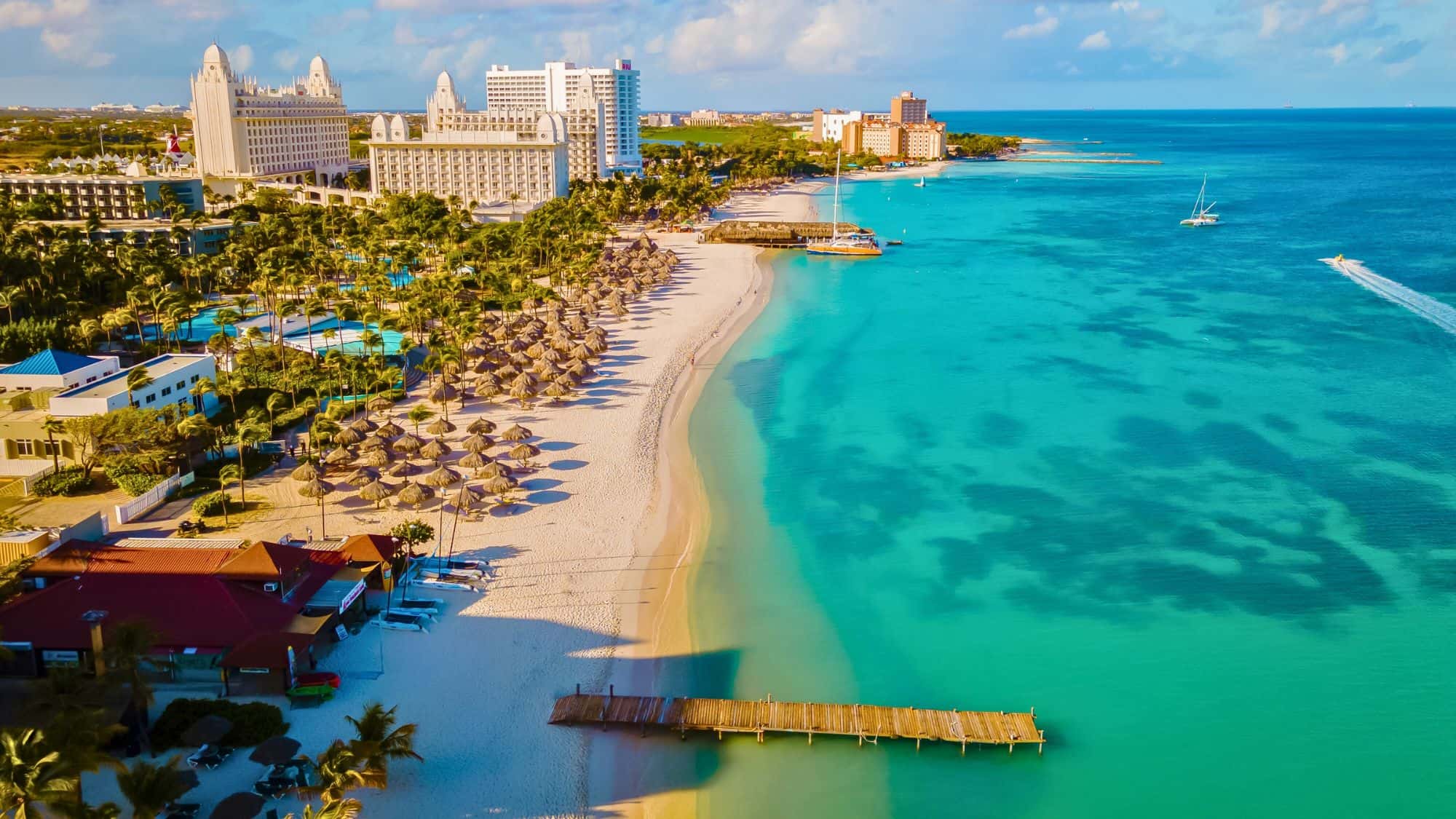 A luxurious tropical beach resort with vibrant turquoise waters, elegant hotels, and beach amenities like thatched umbrellas and loungers. The scene includes palm trees, a wooden pier, and watercraft.
