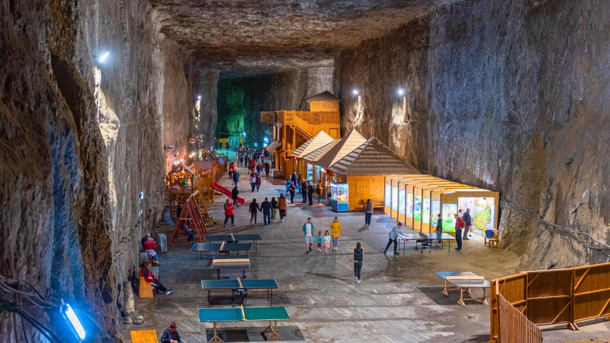 An expansive salt cavern transformed into a recreational space with wooden huts, exhibits, and table tennis, filled with walking visitors.