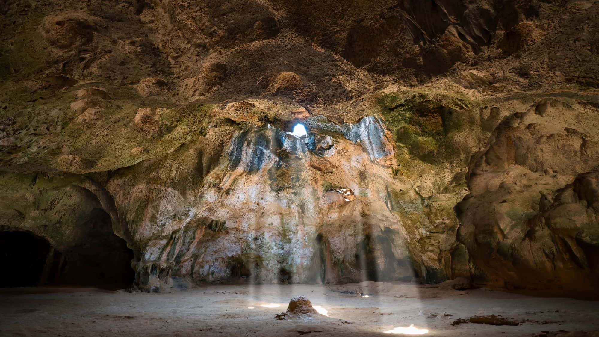 A dramatic cave interior illuminated by sunlight streaming through a small opening. The rugged rock formations and interplay of light and shadow create a picturesque natural setting.