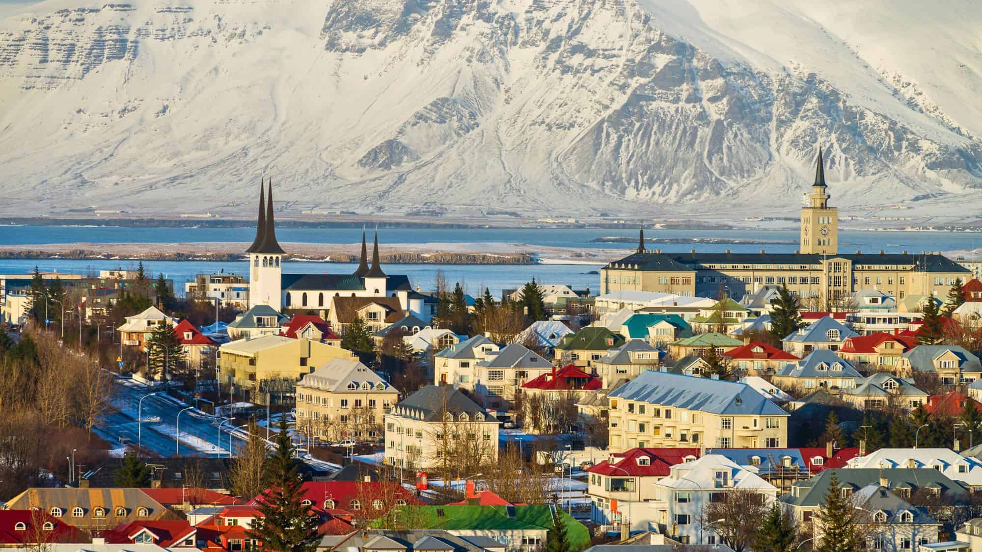 The skyline of Reykjavík features colorful rooftops and pointed church spires with snowy Icelandic mountains towering in the background across a stretch of blue water.