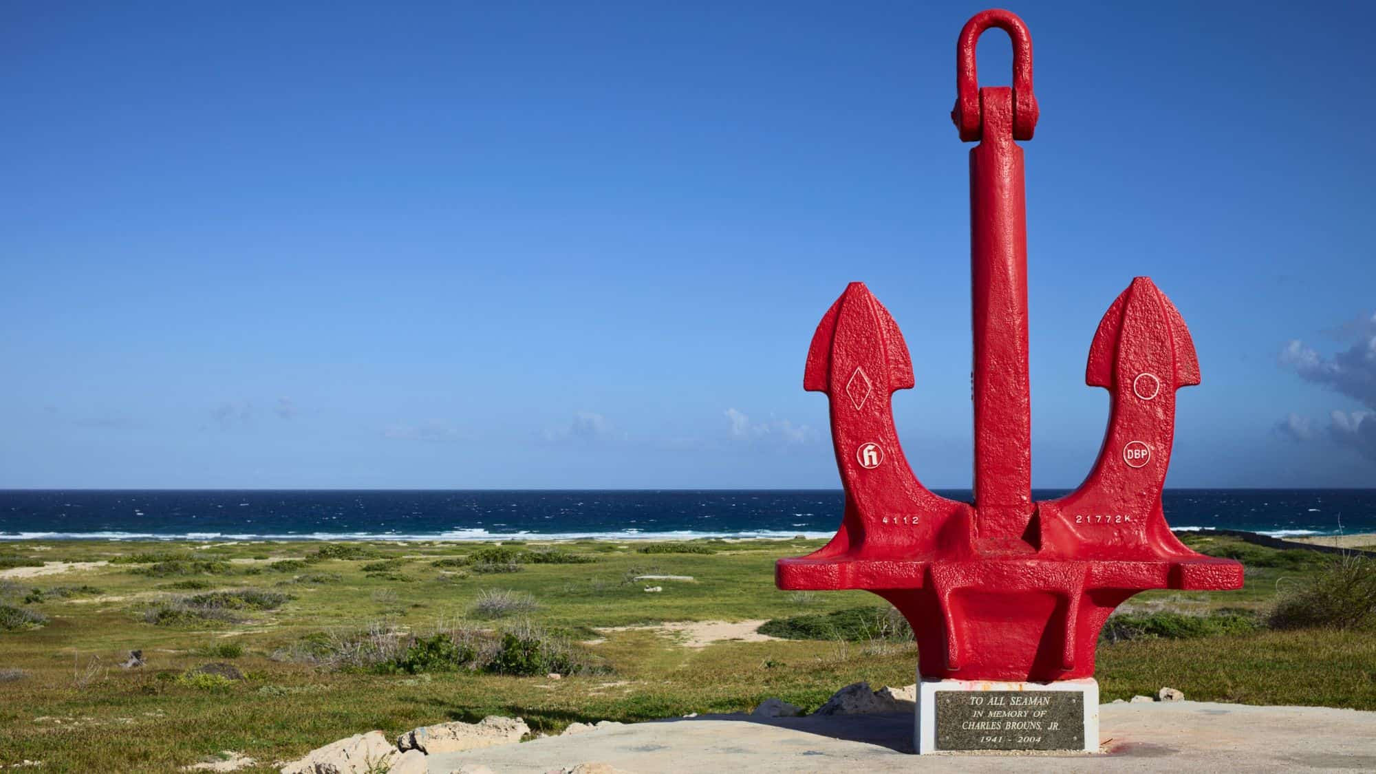 A large red anchor serving as a memorial, set against a serene ocean backdrop. The plaque at the base dedicates it to Charles Brouns, Jr., enhancing its commemorative significance.