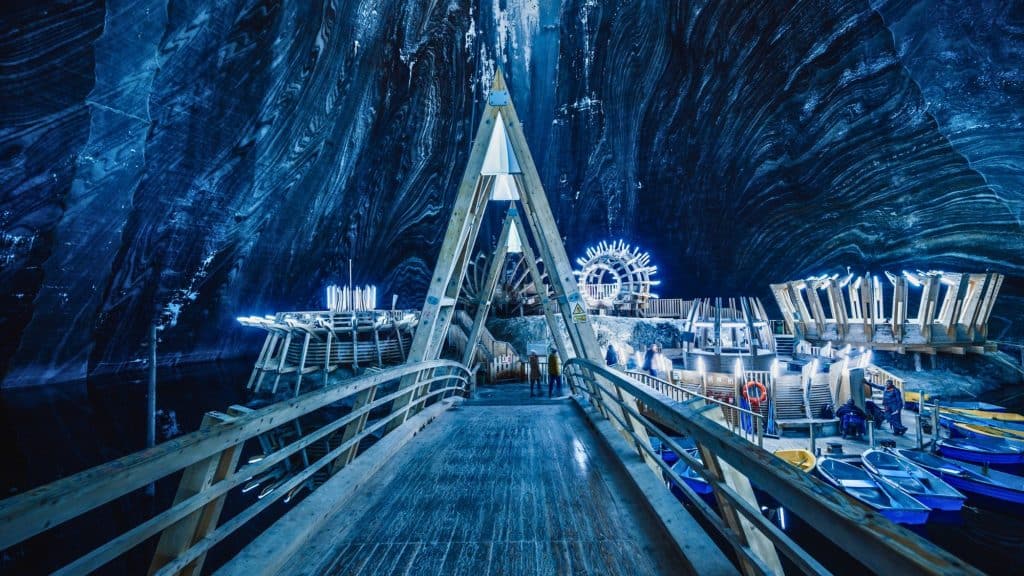 A triangular wooden walkway leads to a sci-fi style dock with glowing installations and small boats inside a massive, striped salt cavern.