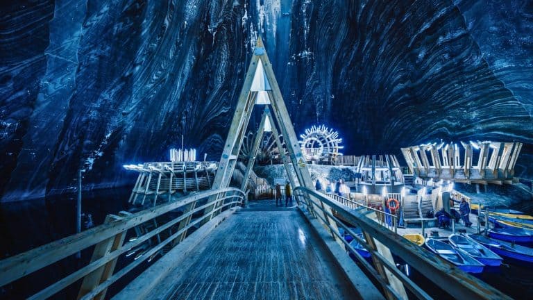 A triangular wooden walkway leads to a sci-fi style dock with glowing installations and small boats inside a massive, striped salt cavern.