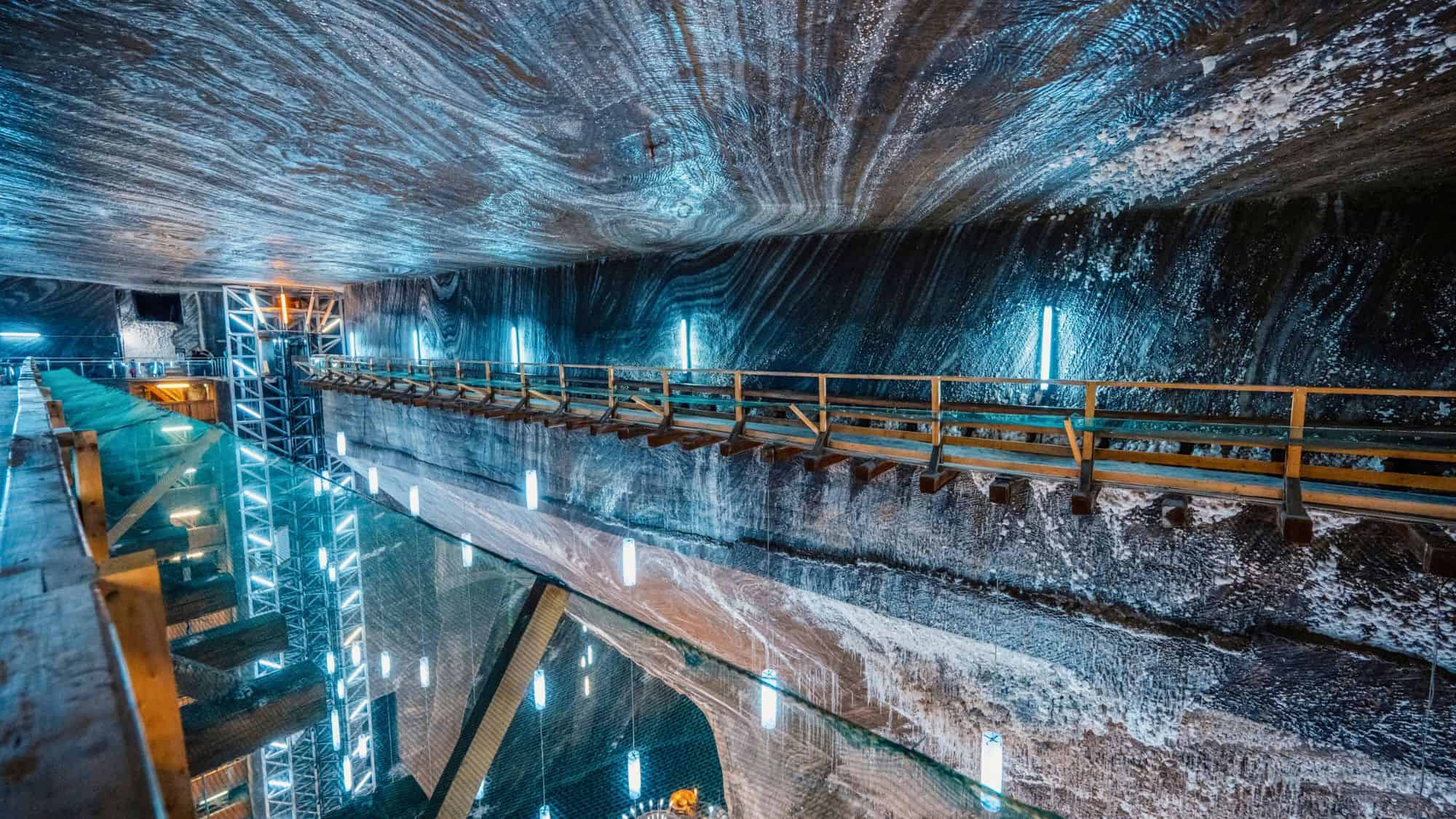 A high wooden walkway runs along the wall of a vast salt chamber with blue lighting and a deep reflective pit beneath.