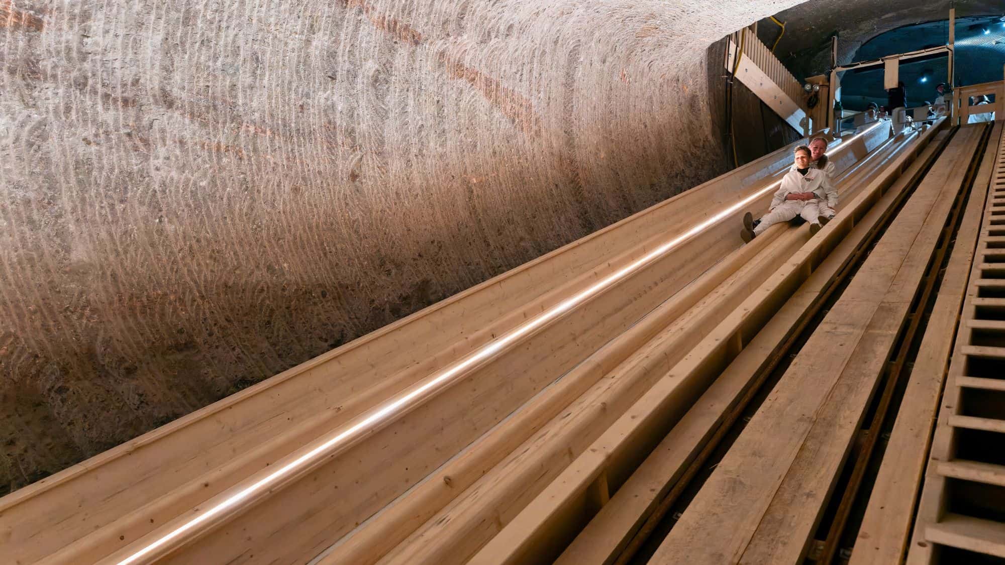 Two people dressed in white slide down parallel wooden chutes carved into a sloped salt tunnel.