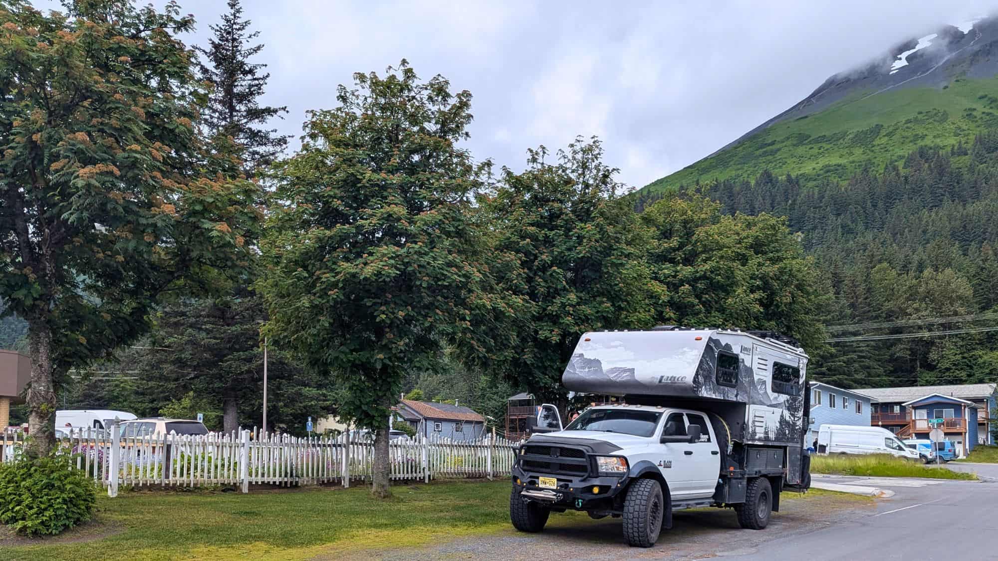 A rugged truck camper is parked on a roadside near white picket fences, modest homes, and lush trees, with a misty mountain slope rising in the background.